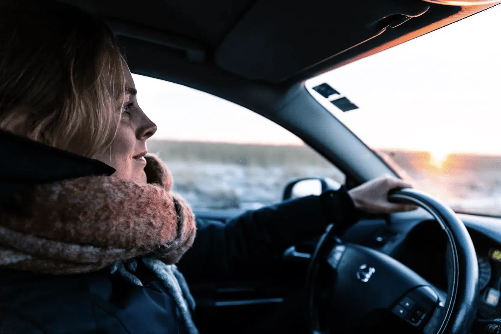 A woman driving a car, dressed warmly in winter clothing, with her hands on the steering wheel. 